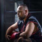 Bearded male boxer in sportswear sitting in boxing ring, contemplating during training session.